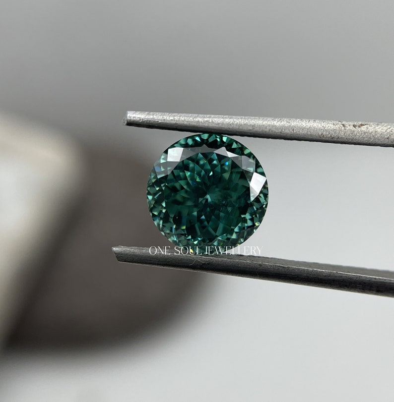 May include: A round, faceted gemstone with a deep green colour. The gemstone is held in tweezers.