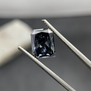 May include: A dark grey emerald cut gemstone held by tweezers. The gemstone has a slight blue hue and is faceted.