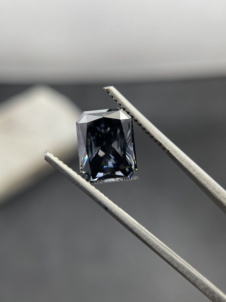 May include: A dark grey emerald cut gemstone held by tweezers. The gemstone has a slight blue hue and is faceted.