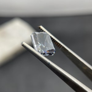 May include: A clear, rectangular, emerald cut gemstone held by tweezers. The gemstone has a slight blue hue and is reflecting light.