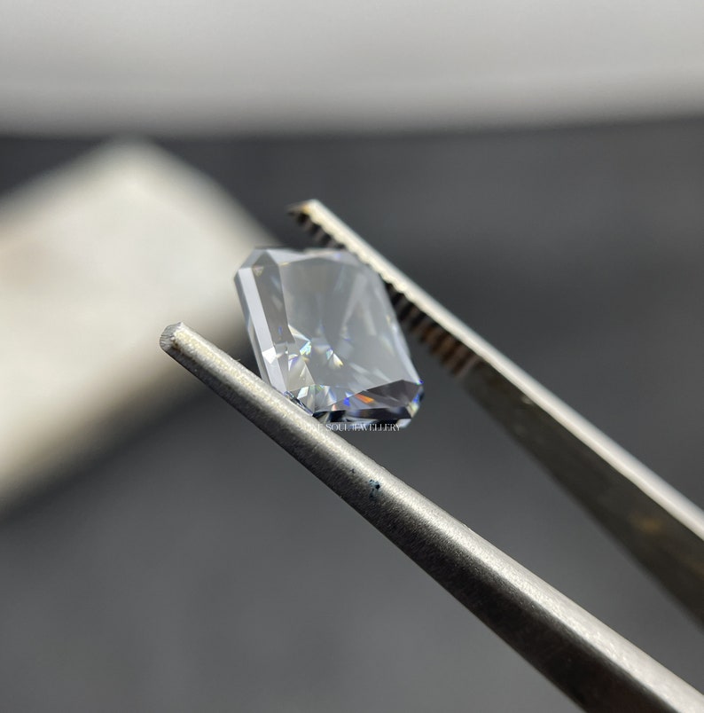 May include: A clear, rectangular, emerald cut gemstone held by tweezers. The gemstone has a slight blue hue and is reflecting light.