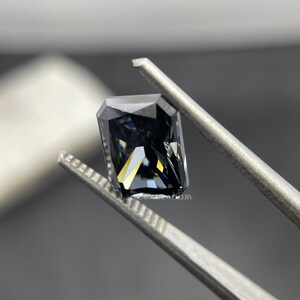 May include: A dark grey, emerald-cut gemstone with a slight blue hue is held by tweezers. The gemstone has a high level of brilliance and sparkle.  ONE SOUL JEWELLERY is printed on the tweezers.