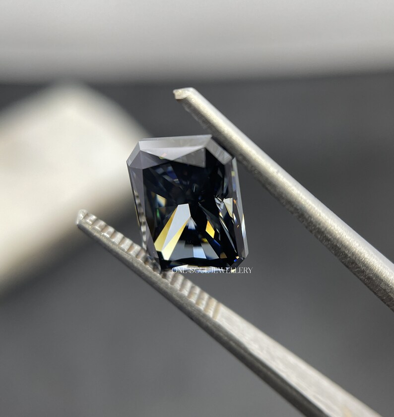 May include: A dark grey, emerald-cut gemstone with a slight blue hue is held by tweezers. The gemstone has a high level of brilliance and sparkle.  ONE SOUL JEWELLERY is printed on the tweezers.