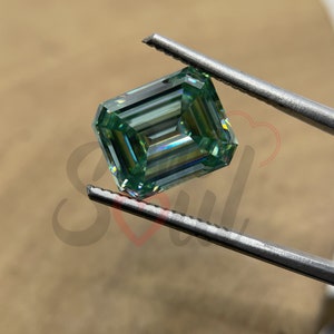 May include: A green emerald cut moissanite gemstone held by tweezers. The gemstone has a brilliant, faceted cut and a light green colour.
