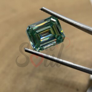 May include: A green emerald cut gemstone with a square shape, held by tweezers. The gemstone has a light green colour and a high level of brilliance.
