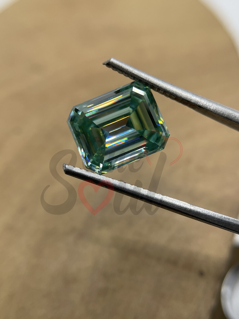 May include: A green emerald cut gemstone with a square shape, held by tweezers. The gemstone has a light green colour and a high level of brilliance.