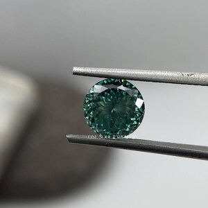 May include: A round, green gemstone with a faceted cut, held by tweezers. The gemstone has a dark green hue with a slight blue tint.