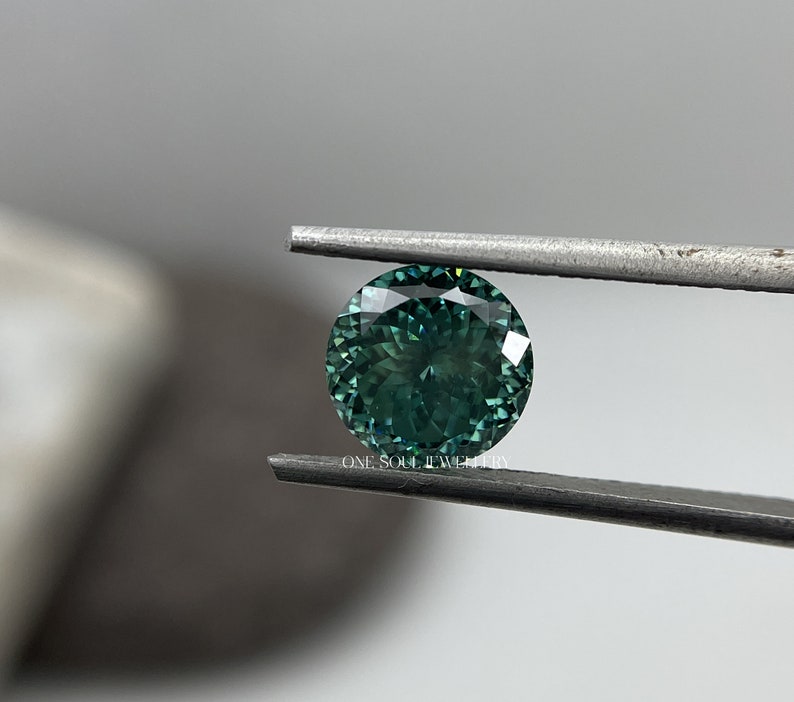May include: A round, green gemstone with a faceted cut, held by tweezers. The gemstone has a dark green hue with a slight blue tint.