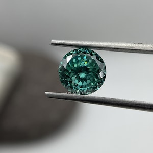 May include: A round, faceted, green gemstone with a slight blue hue. The gemstone is held by tweezers.