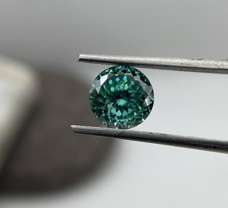 May include: A round, faceted, green gemstone with a slight blue hue. The gemstone is held by tweezers.