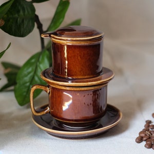 May include: A brown ceramic Vietnamese coffee filter with a lid and a matching cup and saucer.