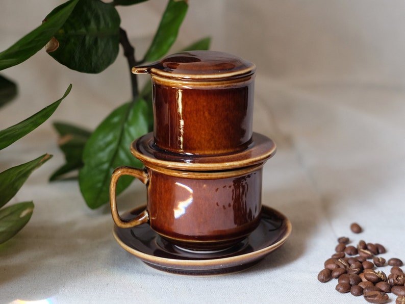 May include: A brown ceramic Vietnamese coffee filter with a lid and a matching cup and saucer.