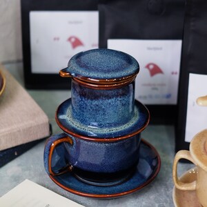 May include: A blue ceramic Vietnamese coffee filter with a lid, a cup, and a saucer. The filter is made of glazed ceramic with a brown rim. The cup and saucer are also blue with a brown rim.
