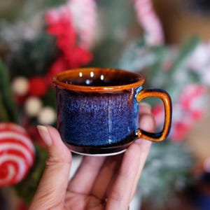 May include: A small ceramic mug with a blue and brown glaze. The mug has a rounded body, a small handle, and a brown rim. The mug is held in a hand, with a blurred background of Christmas decorations.
