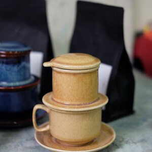 May include: A ceramic coffee filter with a brown and beige color scheme. The filter is stacked on top of a ceramic cup with a saucer. The filter has a lid and a handle. The cup and saucer are also brown and beige.