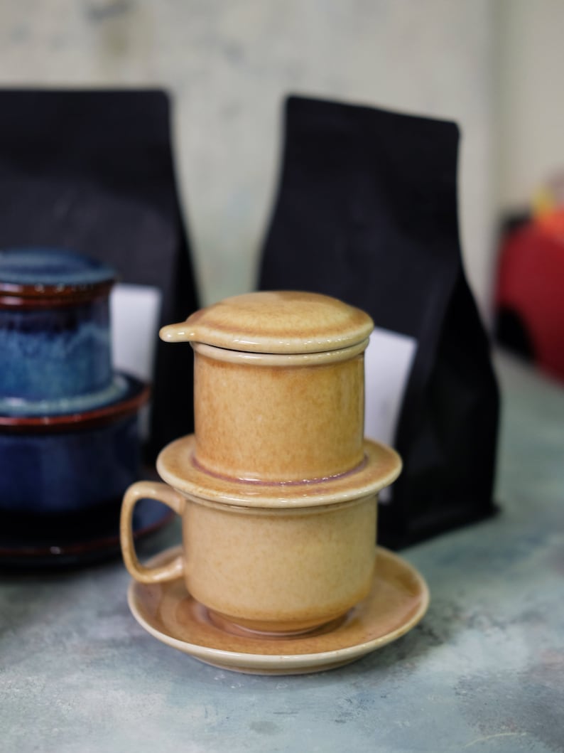 May include: A ceramic coffee filter with a brown and beige color scheme. The filter is stacked on top of a ceramic cup with a saucer. The filter has a lid and a handle. The cup and saucer are also brown and beige.