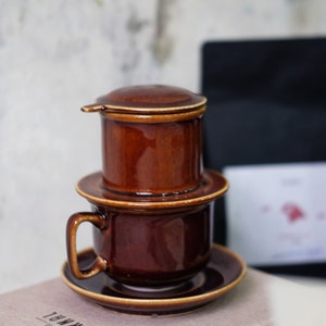 May include: A brown ceramic coffee maker with a filter, a cup, and a saucer. The coffee maker has a lid and a drip spout. The cup and saucer are both brown.
