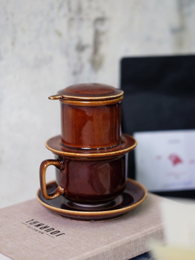 May include: A brown ceramic coffee maker with a filter, a cup, and a saucer. The coffee maker has a lid and a drip spout. The cup and saucer are both brown.