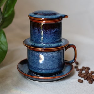 May include: A blue and brown ceramic Vietnamese coffee filter with a drip cup and saucer. The filter is made of two pieces, a top and a bottom, and the drip cup has a handle. There are coffee beans scattered on the table.