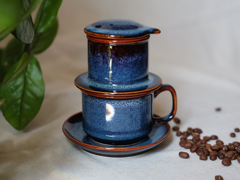 May include: A blue and brown ceramic Vietnamese coffee filter with a drip cup and saucer. The filter is made of two pieces, a top and a bottom, and the drip cup has a handle. There are coffee beans scattered on the table.