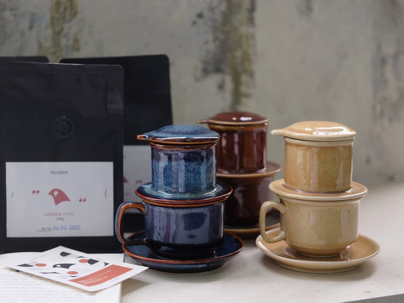 May include: Three ceramic Vietnamese coffee drippers with lids and saucers. The drippers are in blue, brown, and beige. The drippers are stacked on top of each other. The saucers are underneath the drippers. The drippers are on a white surface.