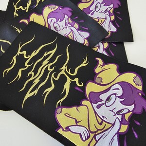 Pu&ograve; includere: Una collezione di toppe rettangolari in tessuto nero con un personaggio dei cartoni animati ricamato con capelli viola, che indossa un mantello giallo con cappuccio e toppe. Il design include linee grafiche gialle a forma di fulmine e accenti viola.