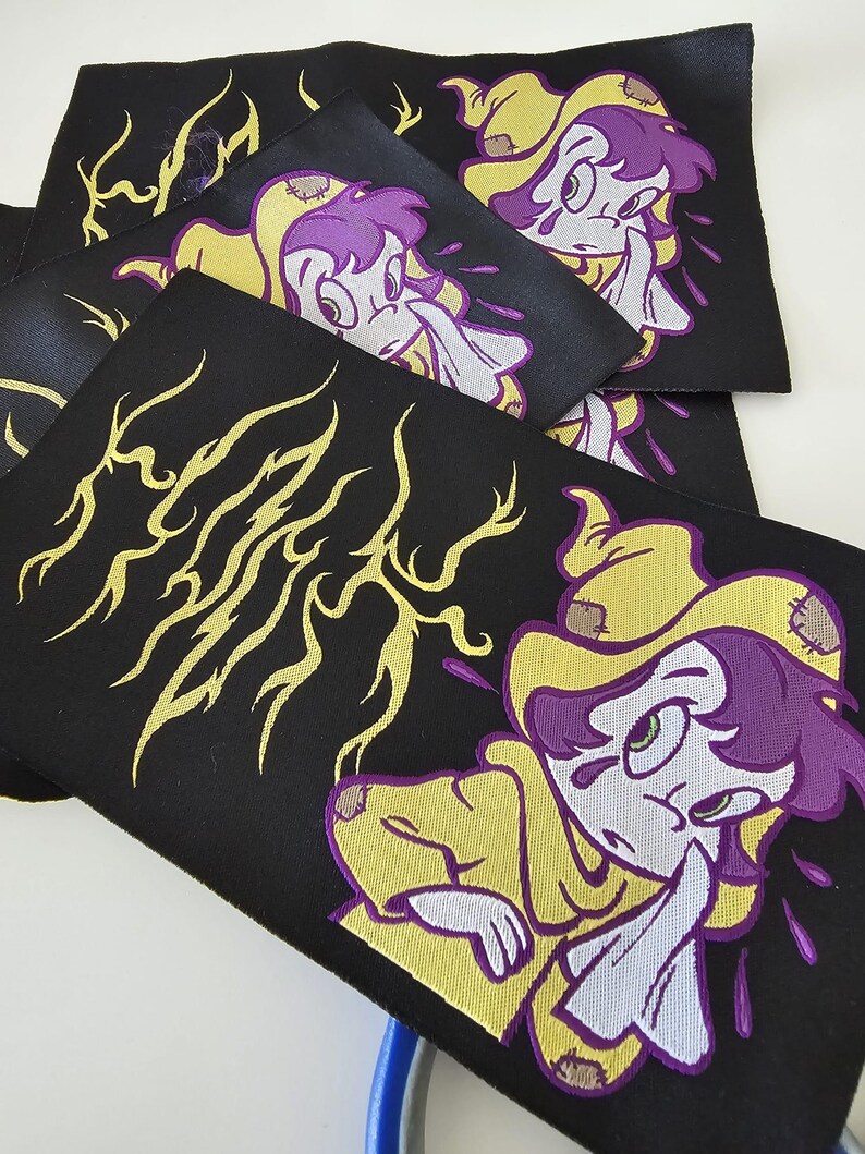 Pu&ograve; includere: Una collezione di toppe rettangolari in tessuto nero con un personaggio dei cartoni animati ricamato con capelli viola, che indossa un mantello giallo con cappuccio e toppe. Il design include linee grafiche gialle a forma di fulmine e accenti viola.
