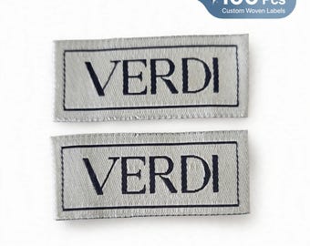 Puede incluir: Dos etiquetas tejidas rectangulares con fondo gris claro, borde oscuro y la palabra VERDI impresa en letras mayúsculas negras. Las etiquetas se presentan en un conjunto de 100 piezas para personalización de marca e identificación de prendas.