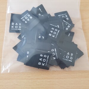 Pu&ograve; includere: Una collezione di piccole etichette in tessuto nero di forma rettangolare all'interno di un sacchetto di plastica trasparente. Ogni etichetta riporta il testo stampato in bianco BAR KOD concept con un piccolo elemento grafico, impilate nella confezione.