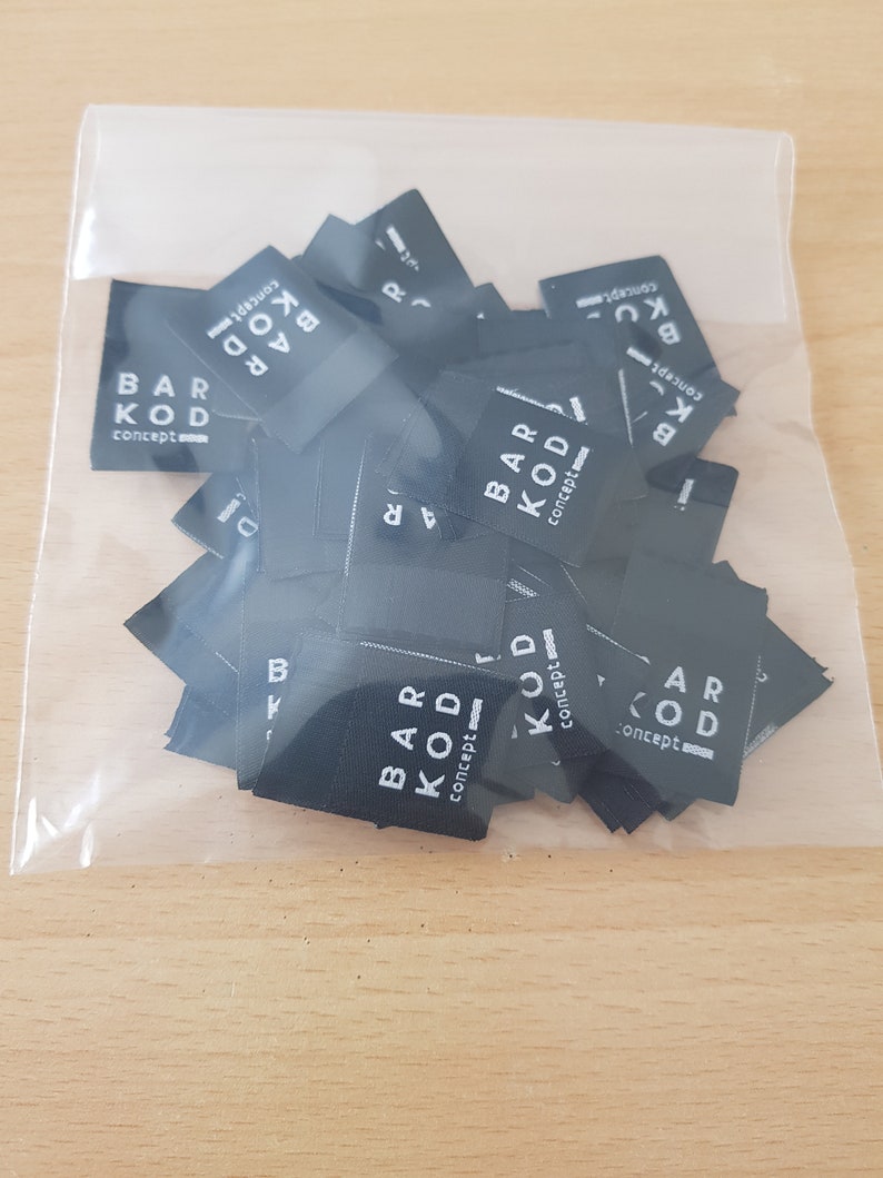 Pu&ograve; includere: Una collezione di piccole etichette in tessuto nero di forma rettangolare all'interno di un sacchetto di plastica trasparente. Ogni etichetta riporta il testo stampato in bianco BAR KOD concept con un piccolo elemento grafico, impilate nella confezione.