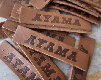 May include: A pile of brown leather tags with the word "AYAMA" embossed in dark brown lettering. The tags are rectangular and appear to be new, with a slightly textured surface. The tags are scattered on a light gray surface.