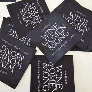 Pu&ograve; includere: Una collezione di etichette quadrate in tessuto nero con testo ricamato in bianco. Ogni etichetta riporta le parole "WINE WOMEN & SONG" in grassetto, con "Look Closer" e "Is On Every Page Of Tha Bible" in caratteri bianchi pi&ugrave; piccoli.