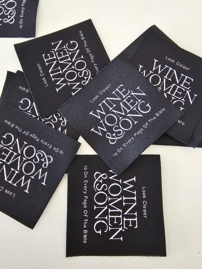 Pu&ograve; includere: Una collezione di etichette quadrate in tessuto nero con testo ricamato in bianco. Ogni etichetta riporta le parole "WINE WOMEN & SONG" in grassetto, con "Look Closer" e "Is On Every Page Of Tha Bible" in caratteri bianchi pi&ugrave; piccoli.