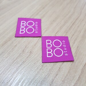 Pu&ograve; includere: Due etichette quadrate in tessuto magenta con il testo bianco BOBO studio stampato in una disposizione sovrapposta. Le etichette presentano una trama tessuta e bordi netti e dritti, mostrando chiaramente il nome del marchio in bianco.