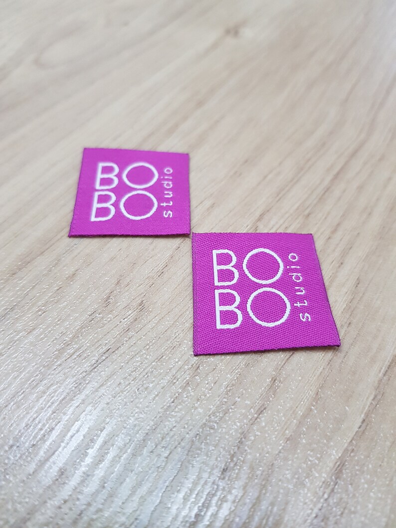 Pu&ograve; includere: Due etichette quadrate in tessuto magenta con il testo bianco BOBO studio stampato in una disposizione sovrapposta. Le etichette presentano una trama tessuta e bordi netti e dritti, mostrando chiaramente il nome del marchio in bianco.
