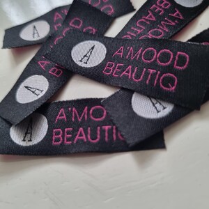 Pu&ograve; includere: Una collezione di etichette in tessuto nero per abbigliamento con un logo circolare bianco contenente una lettera A nera, accompagnato dal testo ricamato in rosa A'MOOD BEAUTIQ. Le etichette rettangolari sono tessute con una finitura strutturata.