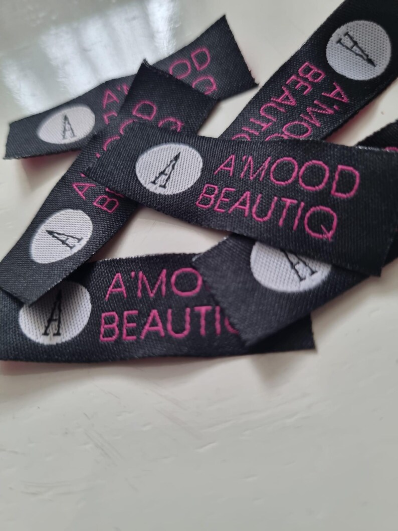 Pu&ograve; includere: Una collezione di etichette in tessuto nero per abbigliamento con un logo circolare bianco contenente una lettera A nera, accompagnato dal testo ricamato in rosa A'MOOD BEAUTIQ. Le etichette rettangolari sono tessute con una finitura strutturata.