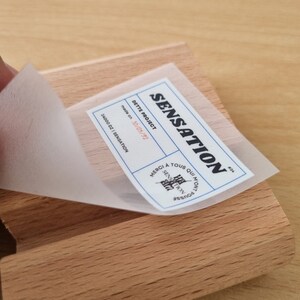 May include: A white sticker with black text that reads "SENSATION DETTE PROJECT made on 30/05/92 24000 DZ/SENSATION" and a small black and white logo that reads "SENSATION" in French. The sticker is on a light brown wooden surface.