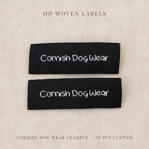 Op de afbeelding: Twee rechthoekige zwarte geweven labels met de wit geborduurde tekst Cornish Dog Wear. De labels zijn horizontaal boven elkaar geplaatst. De tekst HD WOVEN LABELS staat bovenaan en CORNISH DOG WEAR EXAMPLE - 100 PCS CUSTOM staat onderaan.