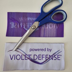Pu&ograve; includere: Un paio di forbici con manici blu e lame in metallo argentato appoggiate su due etichette rettangolari in tessuto. Un'etichetta &egrave; viola con testo bianco, l'altra &egrave; bianca con il testo viola "powered by VIOLET DEFENSE" e un grafico a onde.