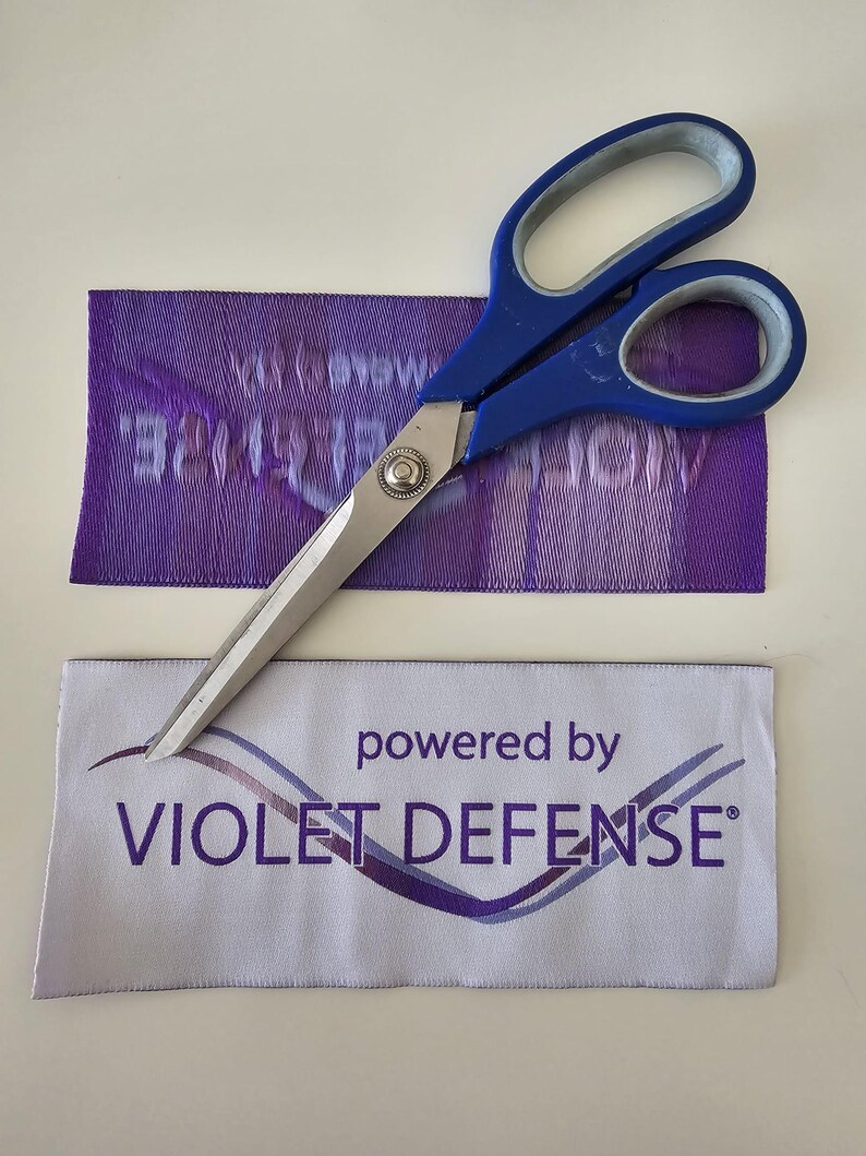 Pu&ograve; includere: Un paio di forbici con manici blu e lame in metallo argentato appoggiate su due etichette rettangolari in tessuto. Un'etichetta &egrave; viola con testo bianco, l'altra &egrave; bianca con il testo viola "powered by VIOLET DEFENSE" e un grafico a onde.