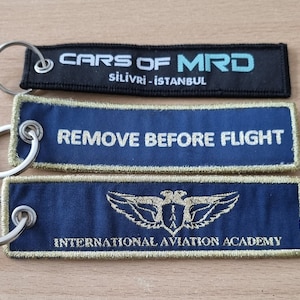 May include: Three keychains with different designs. The top keychain is black with white text that reads "CARS OF MRD" and "silivri - istanbul". The middle keychain is blue with white text that reads "REMOVE BEFORE FLIGHT". The bottom keychain is blue with gold text that reads "INTERNATIONAL AVIATION ACADEMY" and a gold eagle logo.