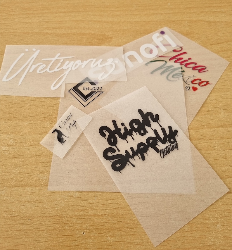 May include: Five iron-on heat transfer vinyl designs on clear sheets. The designs include "High Supply Clothing", "Cavini Pup", "&Uuml;retiyoruz", "znofi", and "Me & Chica Co."
