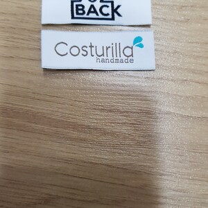 Pu&ograve; includere: Due etichette rettangolari in tessuto bianco per abbigliamento. L'etichetta superiore presenta il testo nero 90Z BACK all'interno di un bordo rettangolare. L'etichetta inferiore mostra il testo marrone Costurilla con la parola handmade sotto e un'icona a forma di goccia d'acqua color verde acqua.