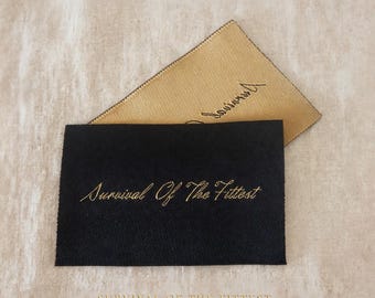 Puede incluir: Dos etiquetas tejidas HD rectangulares, una negra y otra dorada, con el texto Survival Of The Fittest en bordado cursivo dorado. Las etiquetas se muestran con el texto HD WOVEN LABELS en la parte superior y SURVIVAL OF THE FITTEST EXAMPLE - 100 PCS CUSTOM en la parte inferior.
