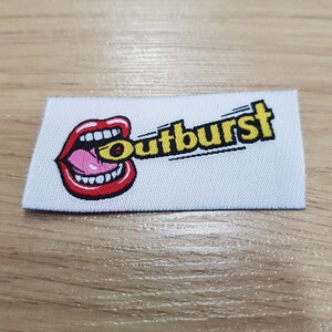 Pu&ograve; includere: Una toppa rettangolare bianca in tessuto con il ricamo di una bocca aperta con labbra rosse, denti bianchi e una lingua rosa. La parola "outburst" &egrave; ricamata in giallo con un contorno nero sulla destra rispetto al grafico della bocca.