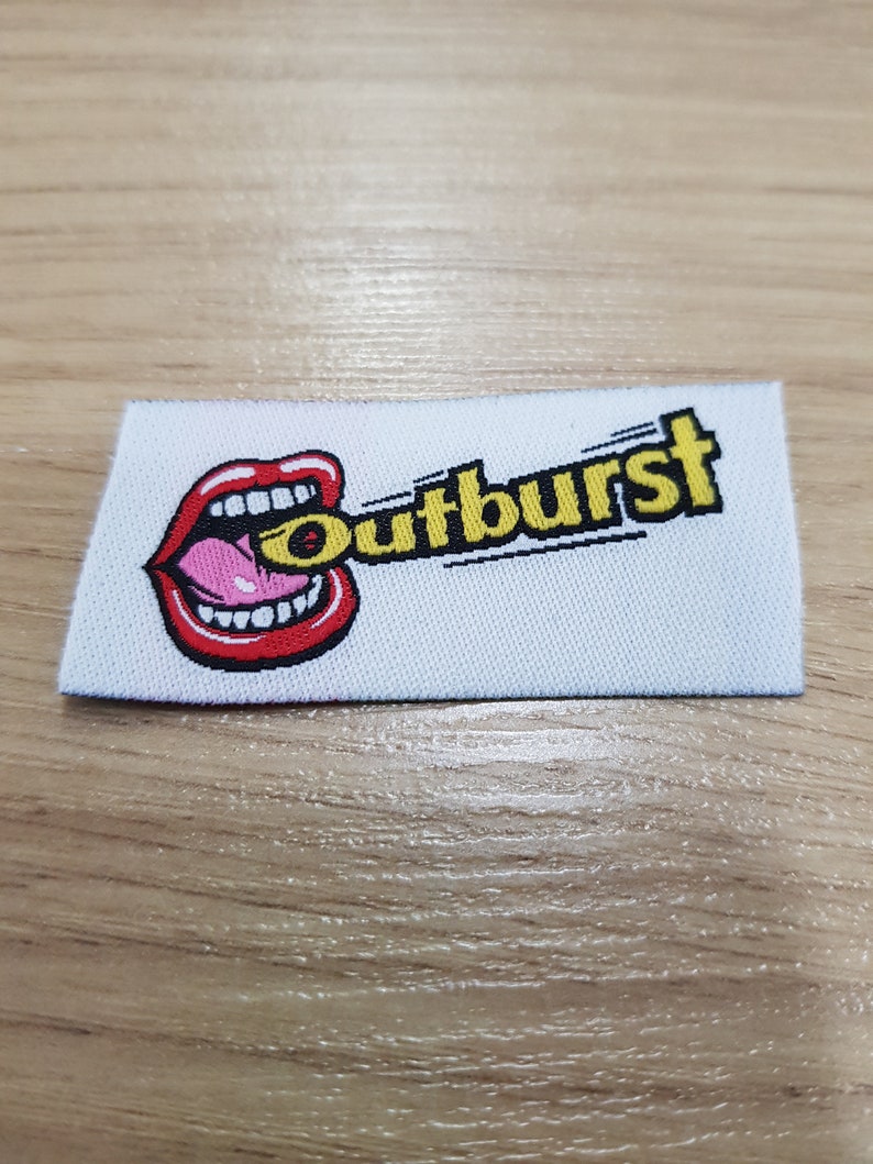 Pu&ograve; includere: Una toppa rettangolare bianca in tessuto con il ricamo di una bocca aperta con labbra rosse, denti bianchi e una lingua rosa. La parola "outburst" &egrave; ricamata in giallo con un contorno nero sulla destra rispetto al grafico della bocca.