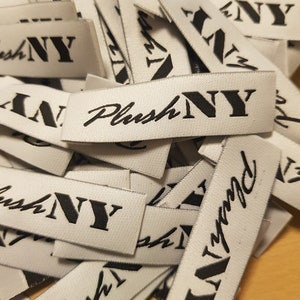 Pu&ograve; includere: Un mucchio di etichette rettangolari in tessuto bianco per abbigliamento con il testo PlushNY ricamato in filo nero. La parola Plush &egrave; scritta in un carattere corsivo, mentre le lettere NY sono visualizzate in un carattere serif grassetto.