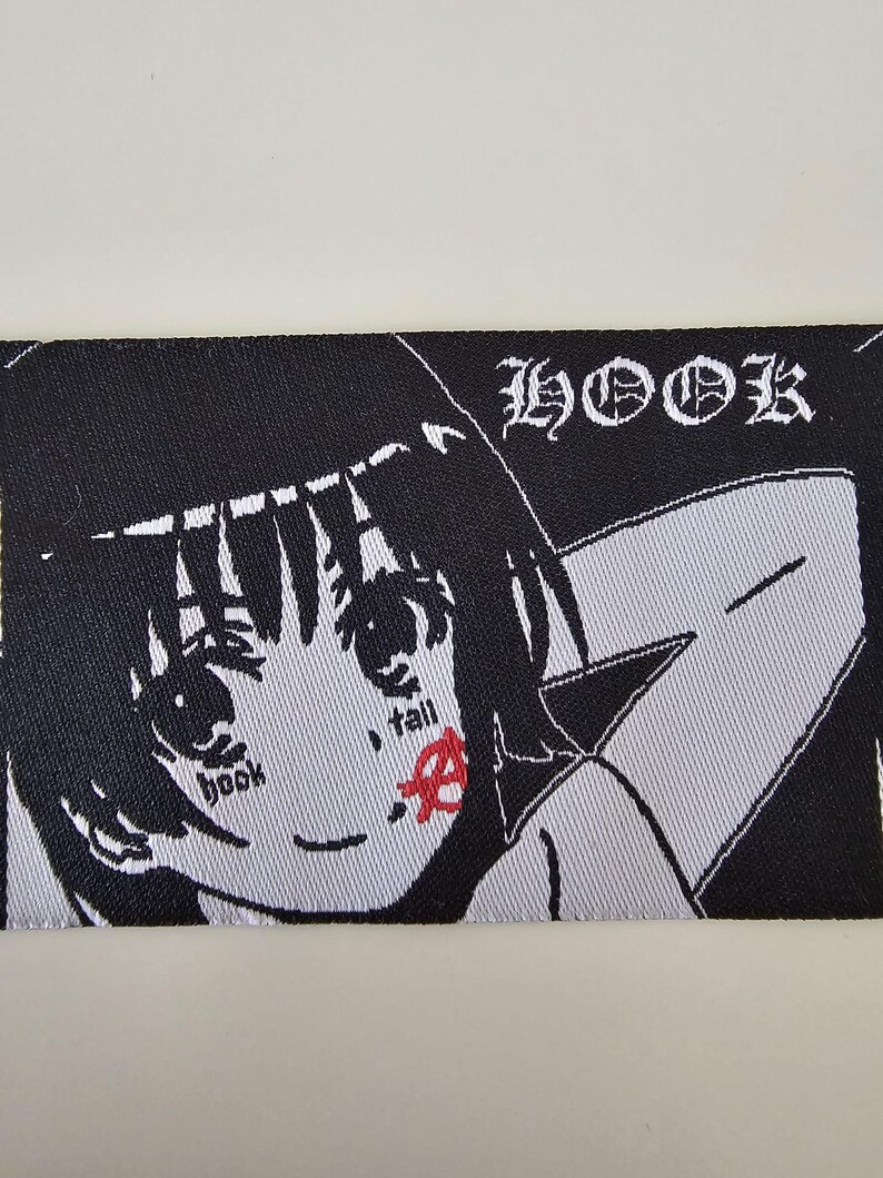 Pu&ograve; includere: Una toppa ricamata rettangolare raffigurante un personaggio in stile anime in bianco e nero con capelli corti. Il testo "HOOK" appare nell'angolo in alto a destra. Il personaggio ha le parole "book" e "fall" sulle guance, con un simbolo anarchico rosso.