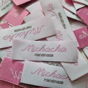 Pu&ograve; includere: Una collezione di etichette rettangolari in tessuto per abbigliamento nei toniamento nei toni del rosa chiaro e del bianco. Ogni etichetta presenta la parola Michacha ricamata in corsivo rosa, con la parola Handmade cucita in filo marrone sotto.